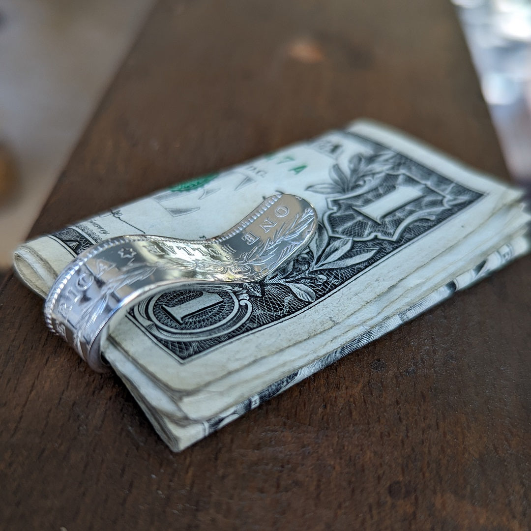 A Morgan Silver Dollar money clip holding a stack of $1 bills, sitting on a wooden bench pin