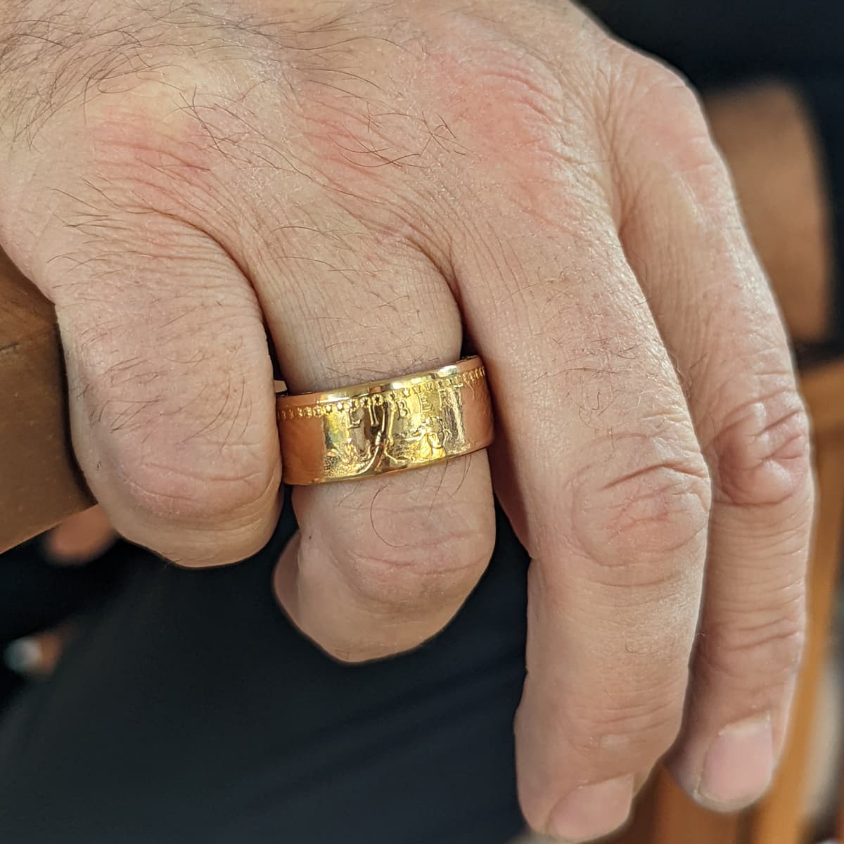 Man's hand wearing an American Gold Eagle coin ring, fingers folded over a wooden railing