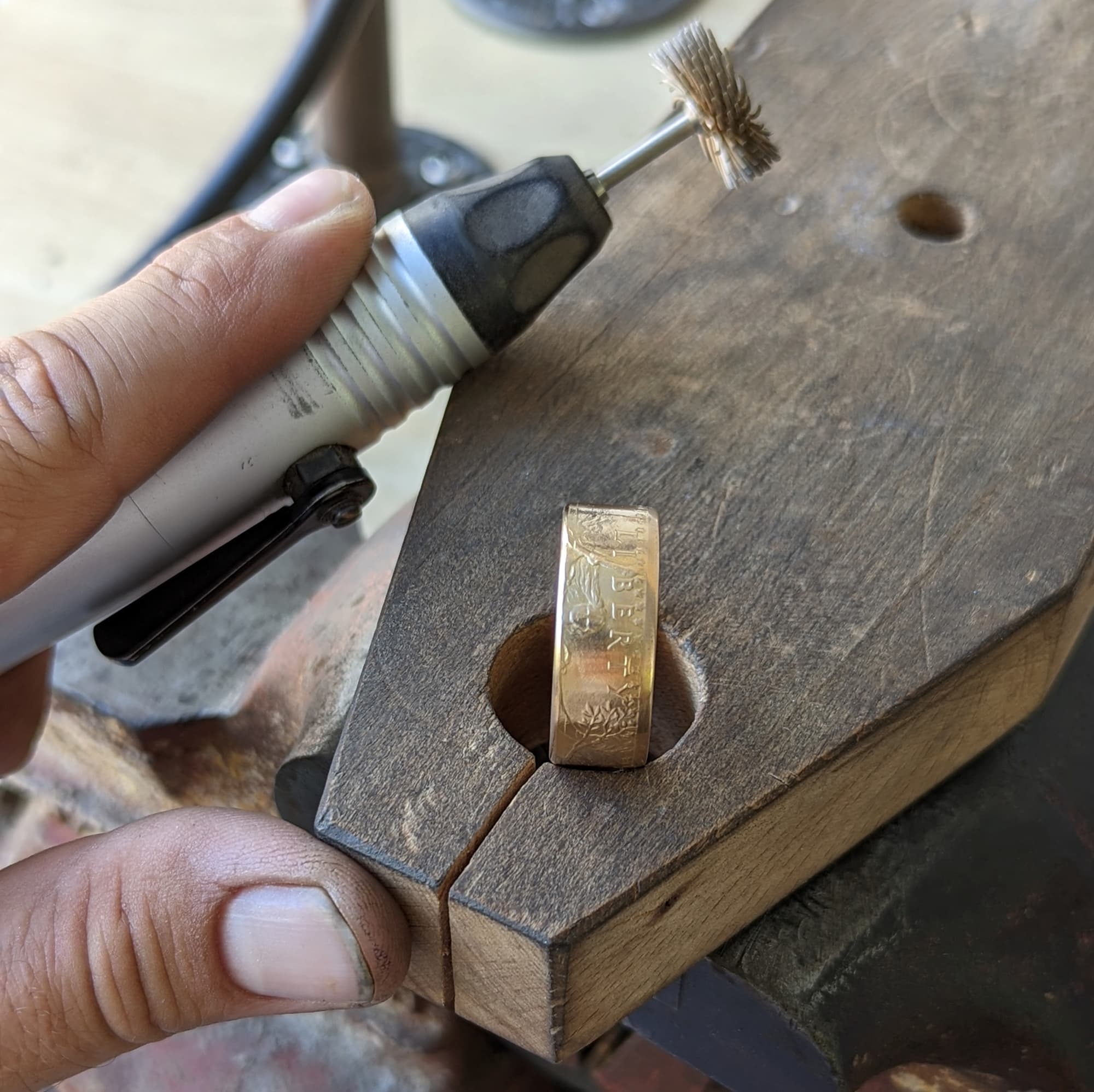 Hand holding a rotary tool, working on a gold coin ring on a wooden bench pin