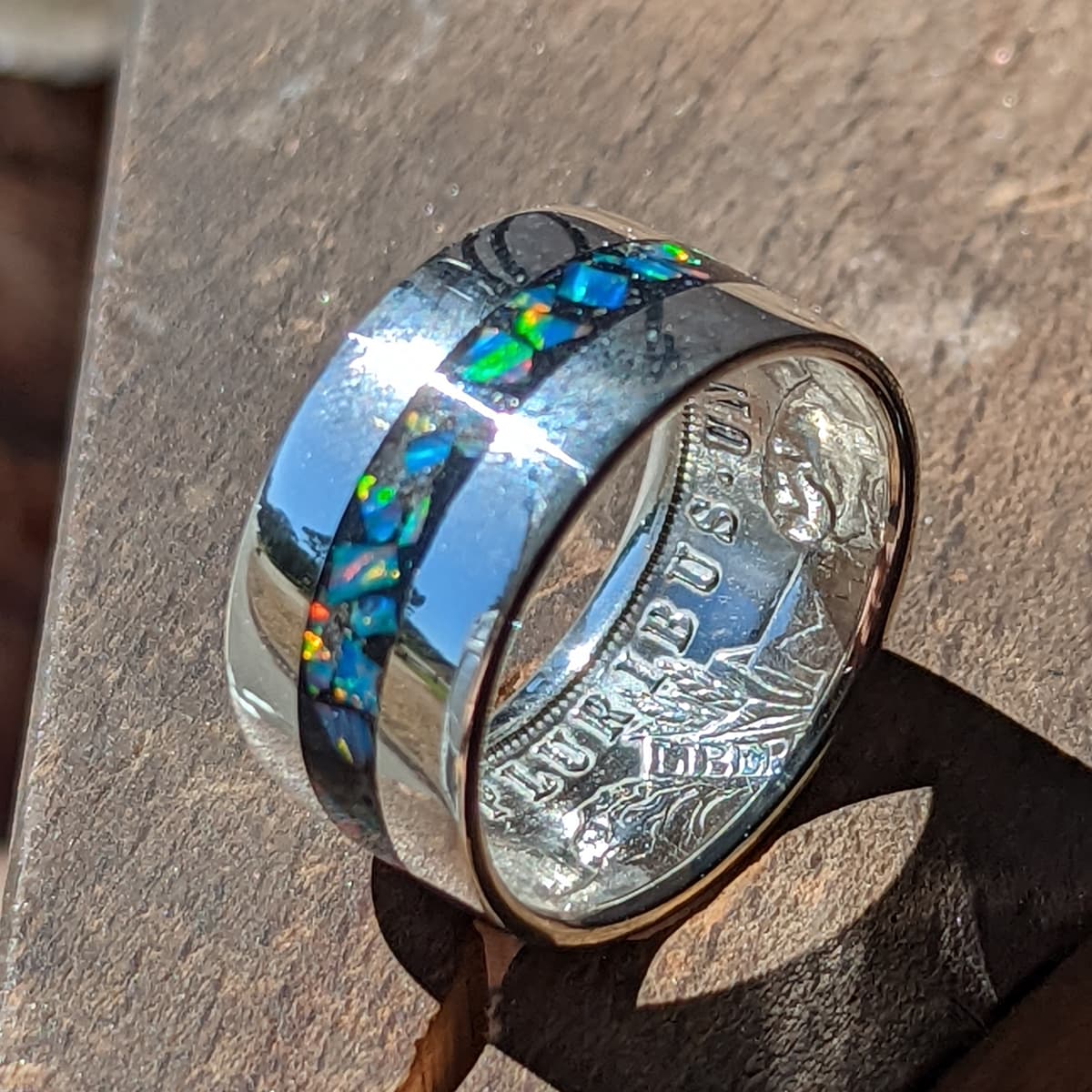 A polished Morgan silver dollar coin ring featuring a central inlay of colorful blue, green, and orange opal stones, photographed close-up on a textured brown wooden surface.