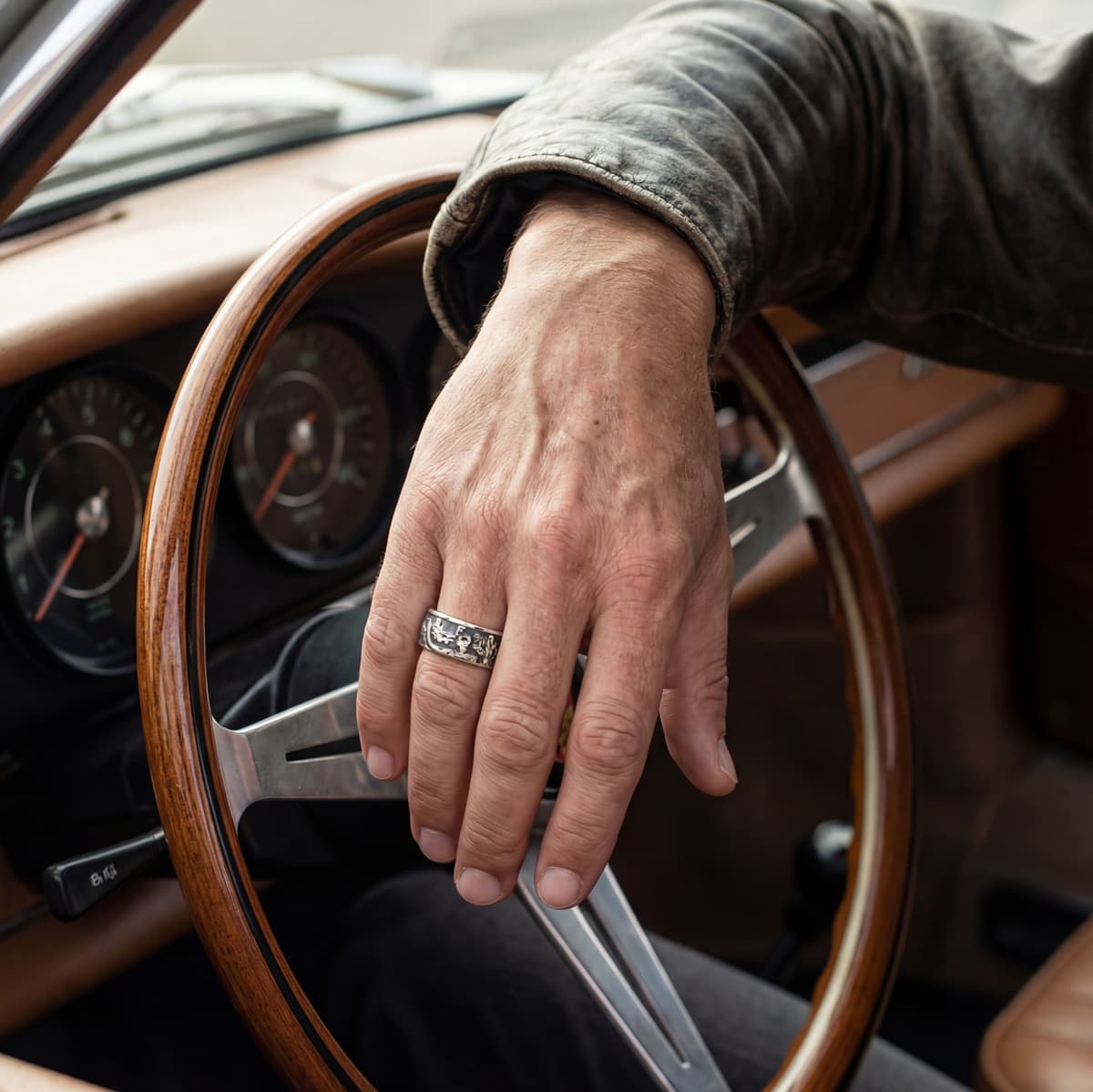 A man wearing a Walking Liberty silver coin ring, is arm resting on the steering wheel of a classic car.